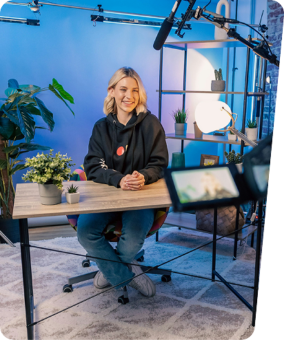 Young woman influencer smiling at desk with plants, camera, and lighting equipment in blue studio environment